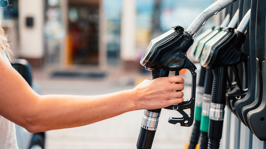 A close-up of a person's arm and hand reaching to grab a fuel pump nozzle at a gas station, with several other nozzles lined up in the background against a blurred storefront.
