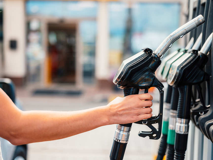 A close-up of a person's arm and hand reaching to grab a fuel pump nozzle at a gas station, with several other nozzles lined up in the background against a blurred storefront.
