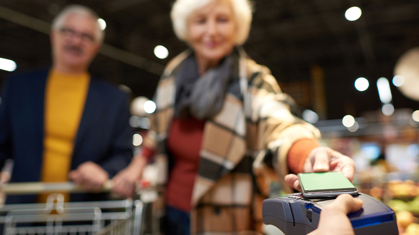 A smiling older woman in a plaid coat holds a smartphone to a card reader to make a contactless payment in a grocery store, while an older man stands behind her with a shopping cart, both softly blurred in the background.