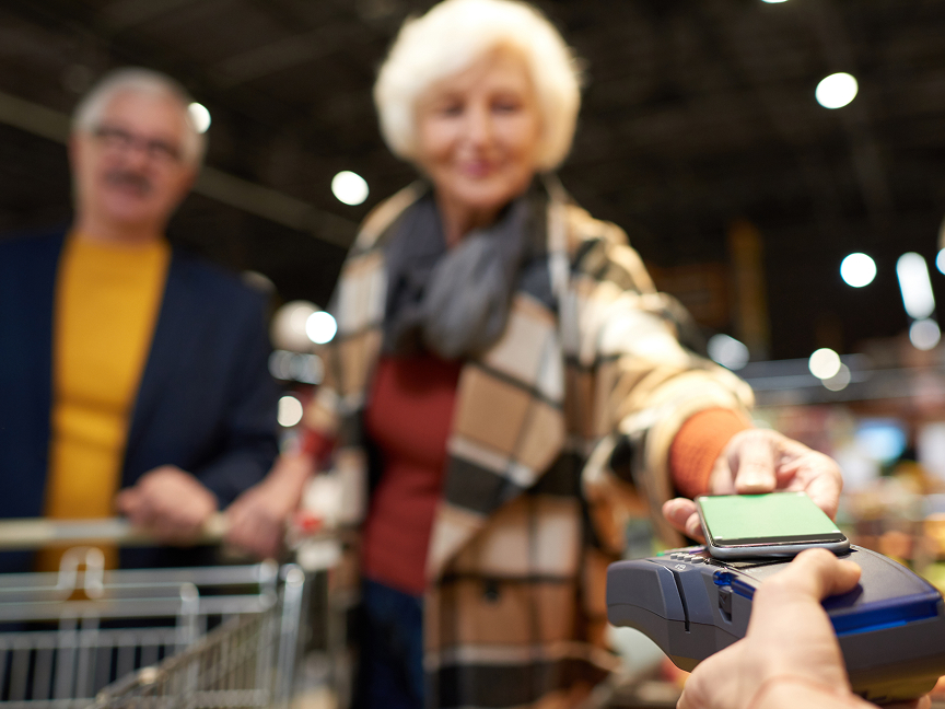 A smiling older woman in a plaid coat holds a smartphone to a card reader to make a contactless payment in a grocery store, while an older man stands behind her with a shopping cart, both softly blurred in the background.