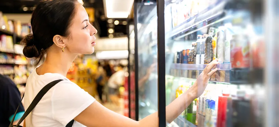Shopper reaching into an open-air cooler for a cold beverage in a convenience store.