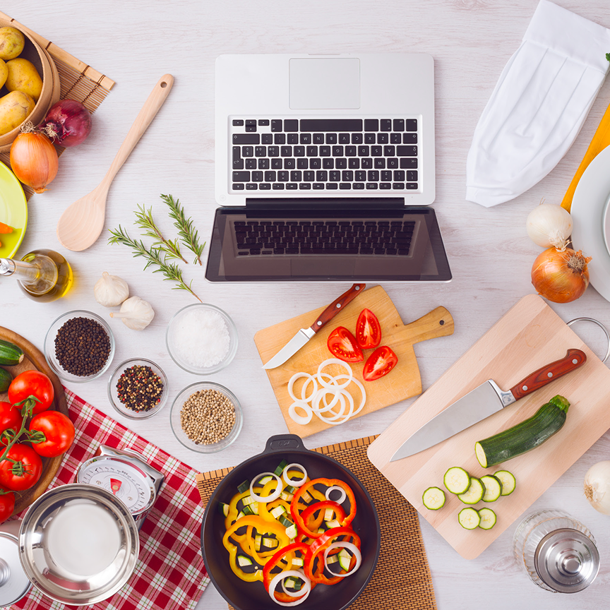 Overhead view of a cooking workspace featuring a laptop at the center, surrounded by fresh vegetables, spices, cutting boards, knives, a chef's hat, and kitchen utensils. Sliced ingredients like zucchini, bell peppers, onions, and tomatoes are arranged neatly, suggesting a on online recipe being followed.