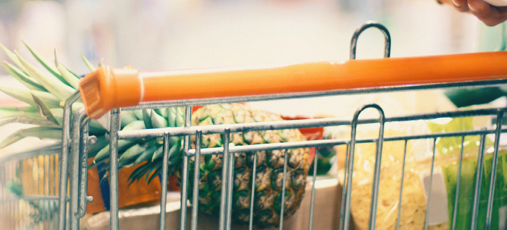 Close-up view of a shopping cart in a store setting, filled with groceries including a whole pineapple, packaged food items, and produce. A person’s hand is visible near the cart handle.