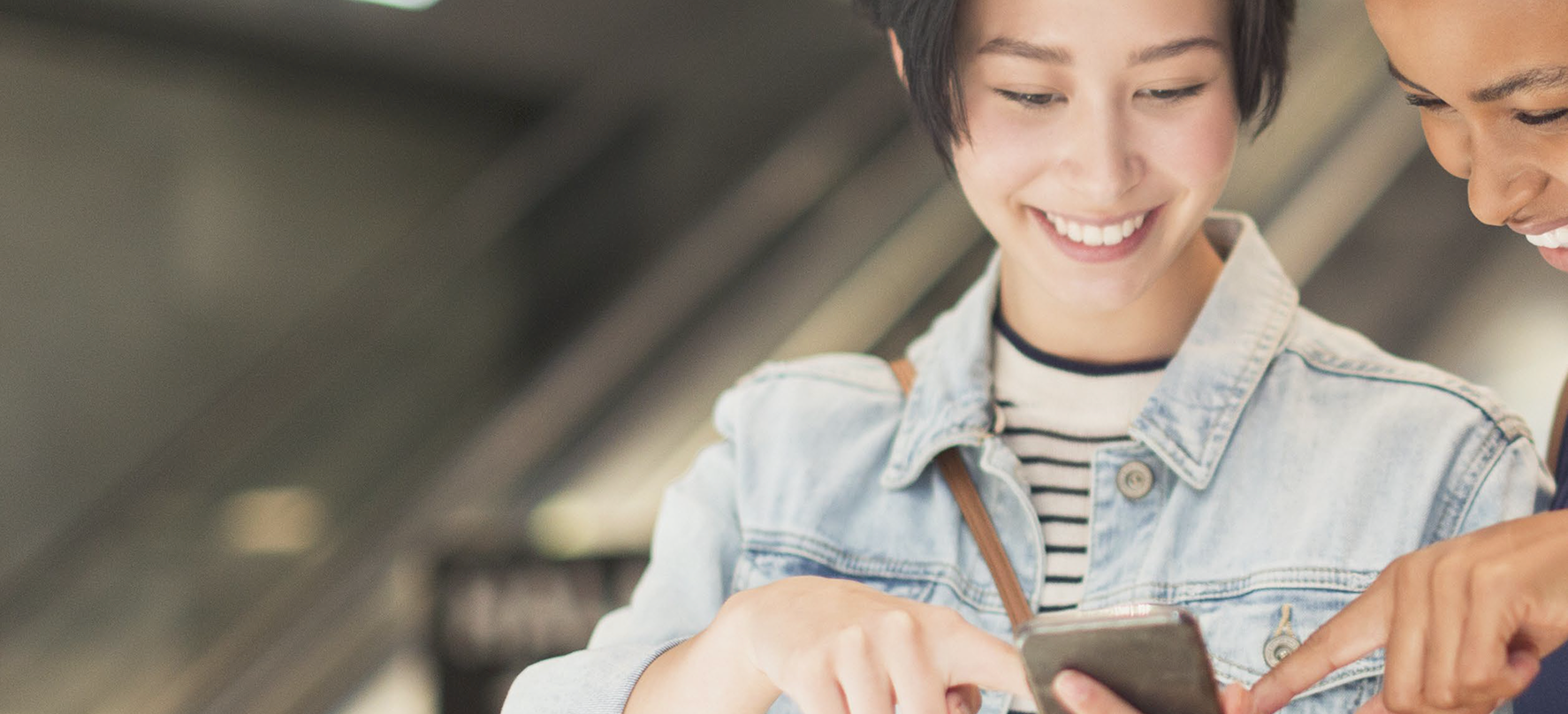 Two smiling individuals looking at a smartphone together. One person is holding the phone, while the other points at the screen. The background shows an indoor setting with escalators, suggesting a shopping mall or retail environment.