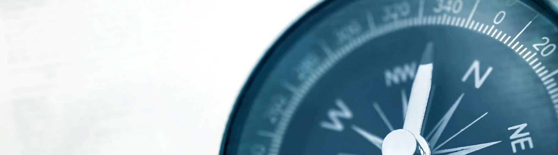 Close-up image of a compass pointing north, with the needle and cardinal directions clearly visible. The background is softly blurred white with hints of text, giving a clean and focused appearance that emphasizes direction and guidance.