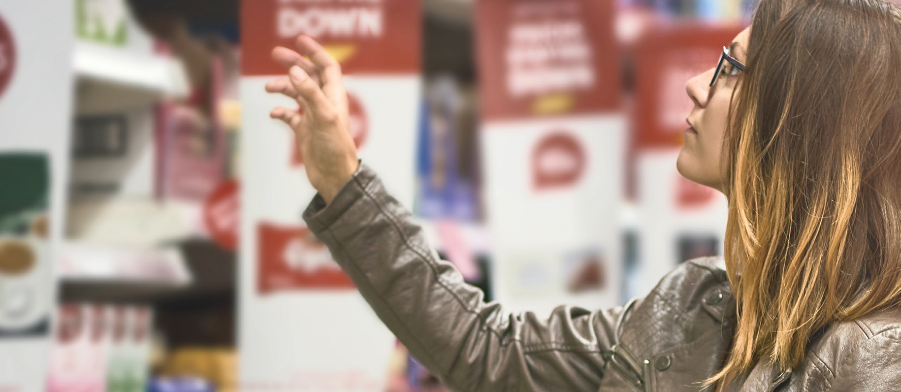 A woman with long hair and glasses, wearing a leather jacket, reaches for a product on a shelf in a brightly lit retail or grocery store.