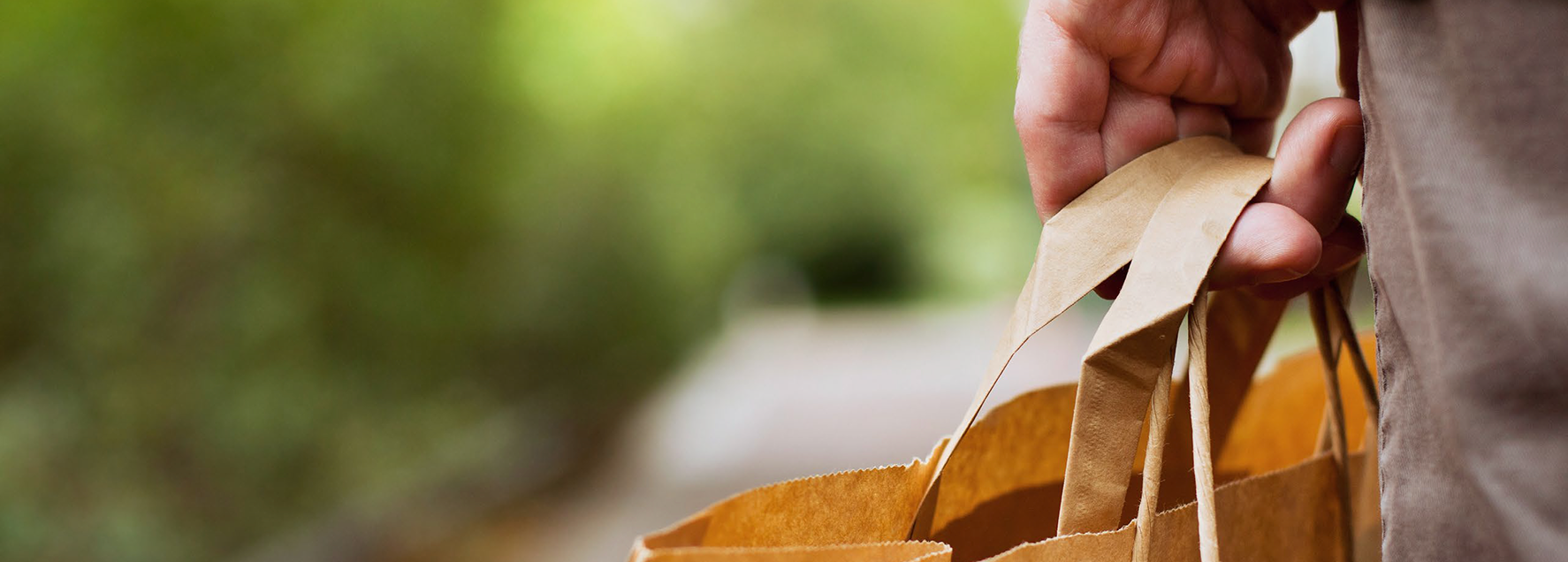 Close-up image of a person’s hand holding the handles of a brown paper shopping bag on a pathway outdoors.