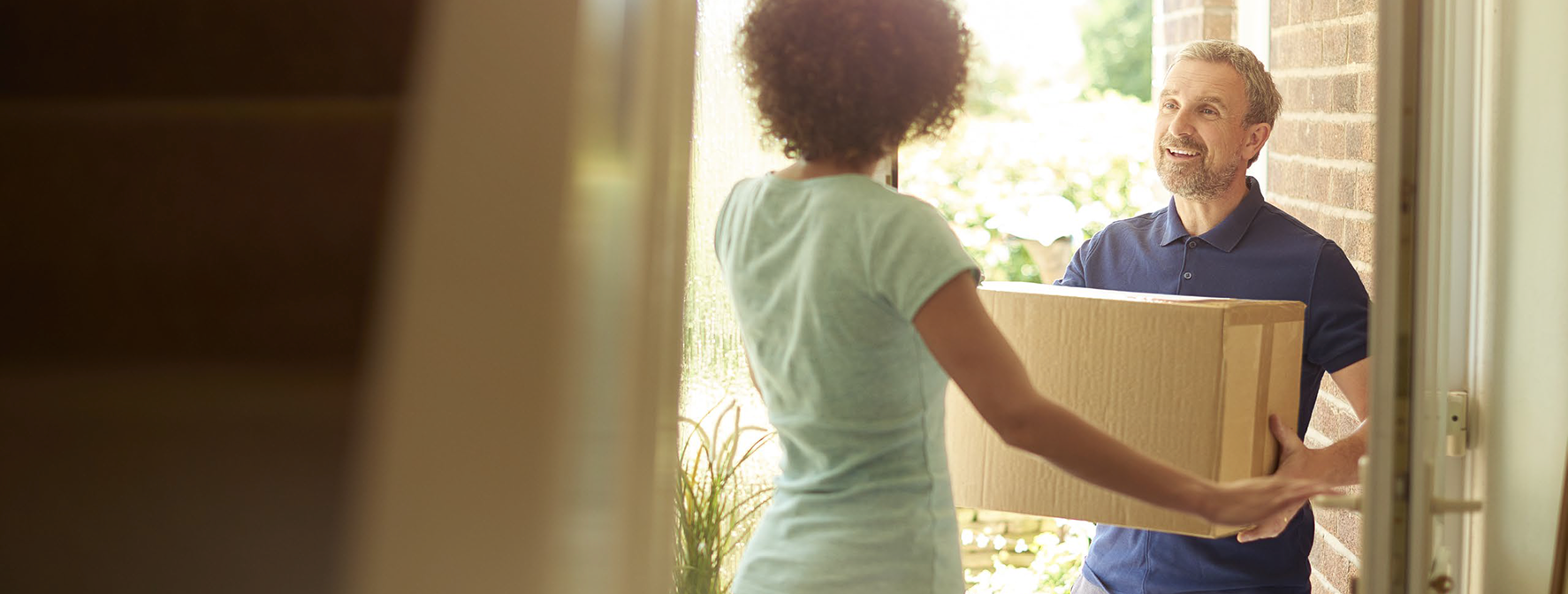 A man stands at the front door of a home smiling as he hands a large cardboard box to a woman who is receiving the package.