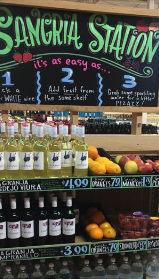 Trader Joe's "Sangria Station” retail display with a colorful chalkboard-style sign reading “It’s as easy as… 1. Pick a red or white wine, 2. Add fruit from the same shelf, 3. Grab some sparkling water for a little pizzazz!” Below the sign, shelves are stocked with bottles of white and red wine, fresh fruit like oranges, mangoes, and strawberries, and bottles of sparkling water, curated for building a DIY sangria.