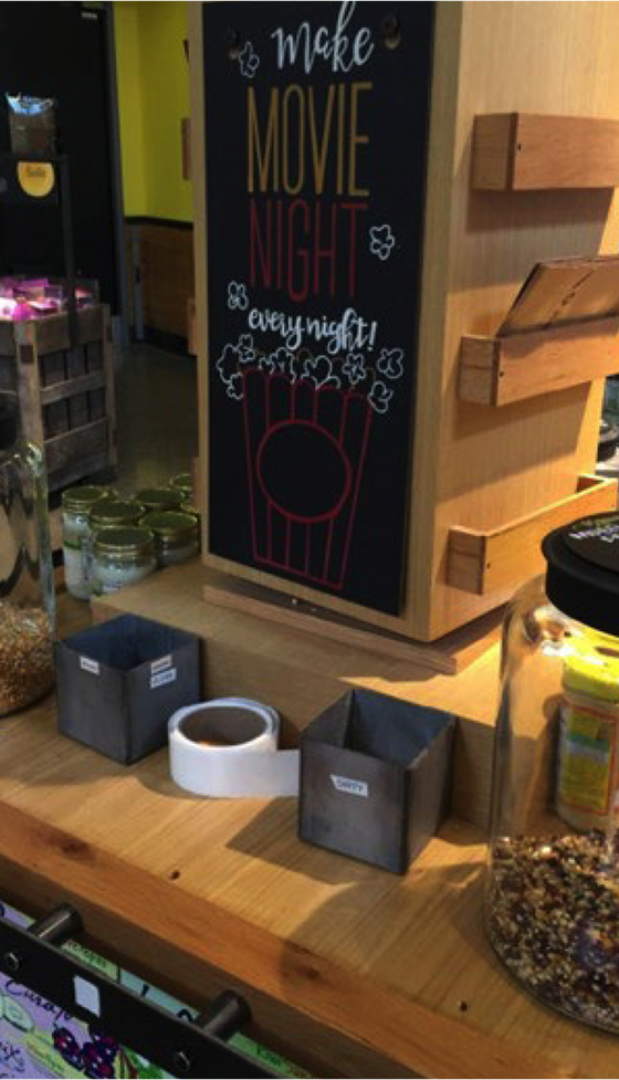 Whole Foods popcorn station display: A hand-drawn chalkboard-style sign reads “Make Movie Night every night!” with popcorn illustrations.