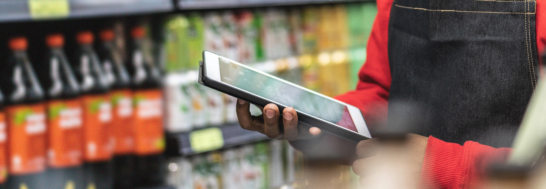 Retail worker using a tablet in front of a shelf stocked with soft drinks and beverages.