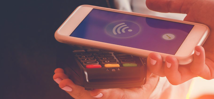 A hand holding a smartphone over a contactless payment terminal, with the screen displaying a wireless payment symbol.
