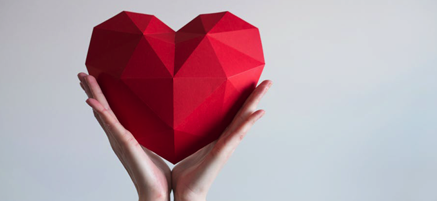 Hands holding a geometric, faceted red heart against a soft gray background.