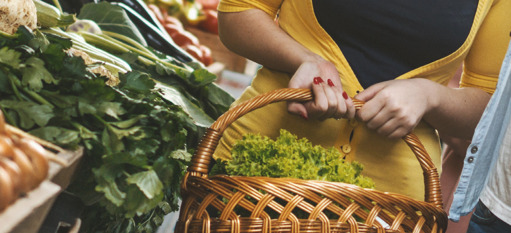 Customer holding a basket with leafy green in a farmer's market setting.