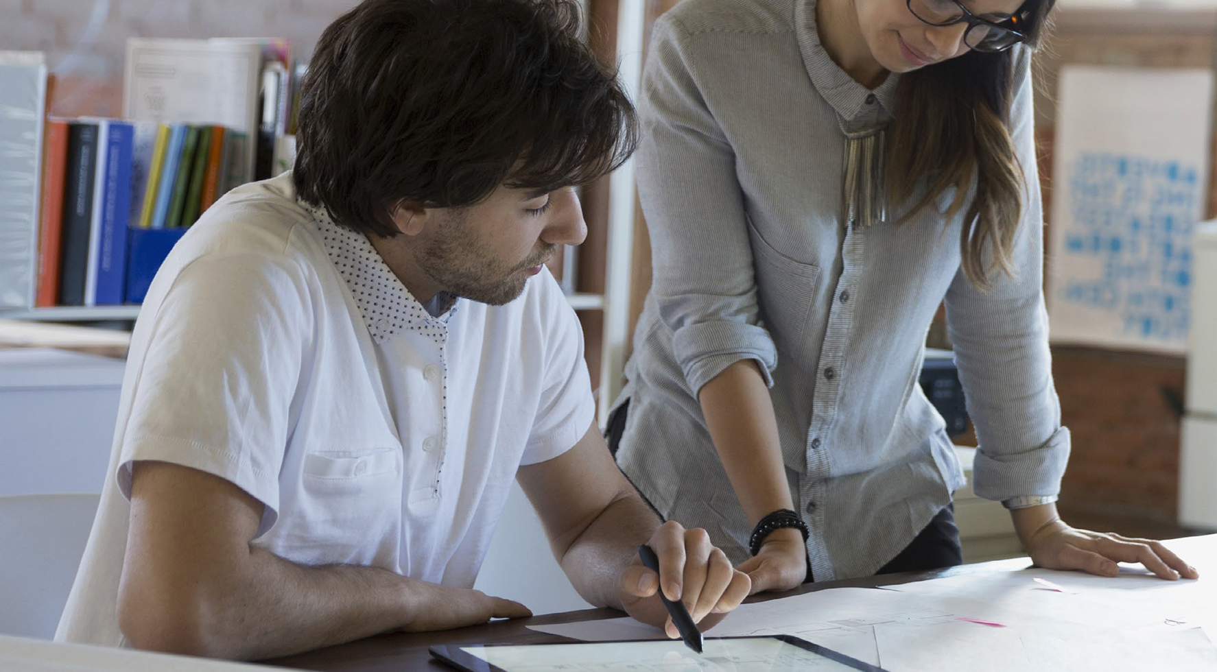Two people collaborating at a desk in an office setting. A man is seated, using a stylus on a tablet, while a woman stands beside him, leaning over a large sheet of paper. They appear to be working on a design or planning project, with shelves of books and office supplies in the background.
