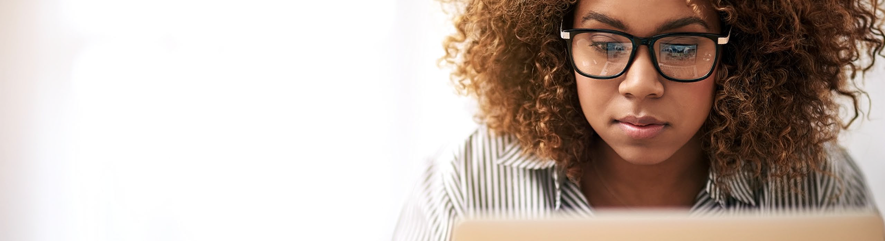 Close-up of a woman with curly hair and glasses focused on her laptop screen in a bright indoor space